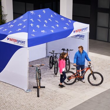 The promotion tent from Intersport is a 4.5x3 m folding gazebo. It is printed all over in white and blue. Various sport icons can be seen on the roof. The salesman shows the customer the bicycle.